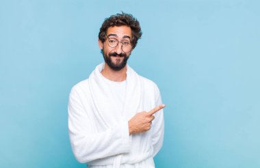 Genç sakallı bir adam banyo cübbesi giyiyor, neşeli bir şekilde gülümsüyor, mutlu hissediyor ve yan ve yukarı doğru işaret ediyor, fotokopi uzayındaki nesneleri gösteriyor.
