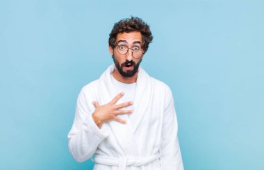 Banyo bornozu giyen genç sakallı adam şaşkın, şaşkın ve şaşırmış hissediyor, elleri göğsünde ve ağzı açık, kim olduğumu söylüyor, ben?