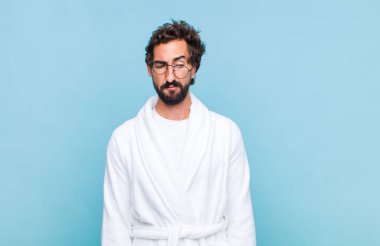 Genç sakallı bir adam banyo cübbesi giyiyor üzgün, üzgün ya da kızgın ve olumsuz bir tavır takınıyor, anlaşmazlık içinde kaşlarını çatıyor.