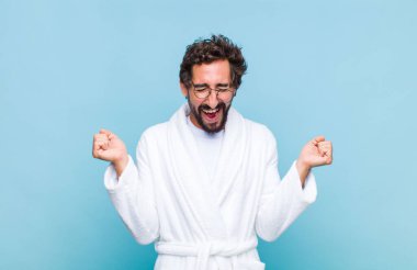 Banyo bornozu giymiş genç sakallı adam son derece mutlu ve şaşırmış görünüyor, başarıyı kutluyor, bağırıyor ve zıplıyor.