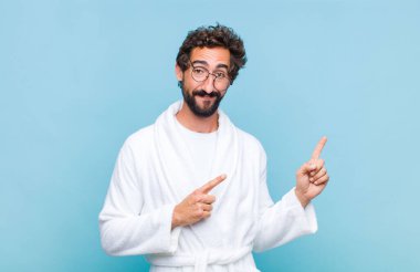 Banyo bornozu giymiş genç sakallı adam mutlu bir şekilde gülümsüyor ve her iki eli de fotokopi uzayında nesne gösteriyor.