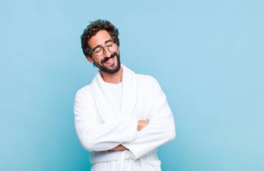 Banyo bornozu giymiş genç sakallı adam kollarını kavuşturup mutlu bir şekilde gülüyor, rahat, pozitif ve tatmin edici bir pozla.