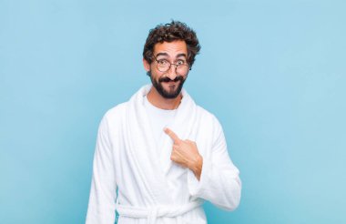 Banyo bornozu giyen genç sakallı adam mutlu, gururlu ve şaşırmış görünüyor, neşeli bir şekilde kendini gösteriyor, kendinden emin ve yüce hissediyor.