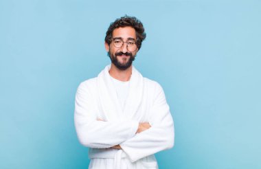 Banyo cübbesi giyen genç sakallı adam mutlu, gururlu ve tatmin olmuş kolları bağlı gülümseyen biri gibi görünüyor.