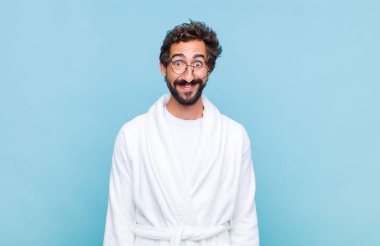 Banyo cübbesi giyen genç sakallı adam neşeli ve şapşal görünüyor, geniş, eğlenceli, kaçık bir gülüşü ve açık gözleri var.