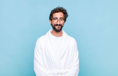 Banyo bornozu giymiş genç sakallı adam olumlu ve kendinden emin bir şekilde gülümsüyor. Tatmin olmuş, arkadaş canlısı ve mutlu görünüyor.