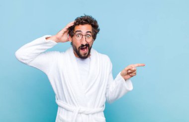 Genç sakallı adam banyo cübbesi giyiyor gülüyor, mutlu, pozitif ve şaşırmış görünüyor, yanal fotokopi alanını işaret eden harika bir fikri fark ediyor.