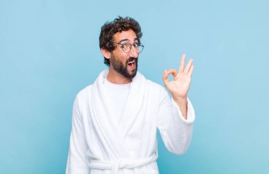 Banyo bornozu giyen genç sakallı adam kendini başarılı ve tatmin olmuş hissediyor, ağzı açık gülümsüyor, eliyle işaret veriyor.