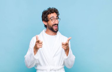 Banyo bornozu giymiş genç sakallı bir adam. Pozitif, başarılı ve mutlu bir tavırla gülümsüyor. Kamerayı işaret ediyor.