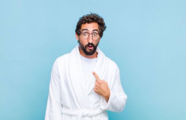 Banyo bornozu giymiş genç sakallı adam ağzı açık, şaşkın ve şaşırmış bir şekilde kendini işaret ediyor.
