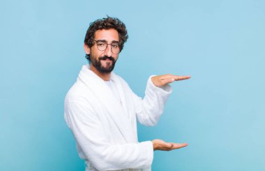 Banyo bornozu giymiş genç sakallı bir adam iki eli yanda bir nesne tutuyor fotokopi alanı, bir nesneyi gösteriyor, sunuyor ya da reklamını yapıyor.