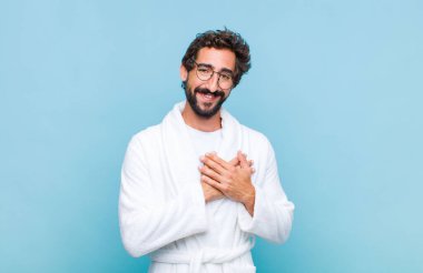 Genç sakallı bir adam bornoz giyiyor. Romantik, mutlu ve aşık. Neşeli bir şekilde gülümsüyor ve kalbe yakın el ele tutuşuyor.