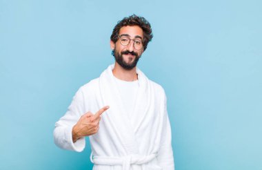 Banyo bornozu giyen genç sakallı adam gururlu, kendinden emin ve mutlu görünüyor, gülümsüyor ve kendini işaret ediyor ya da bir numara işareti yapıyor.