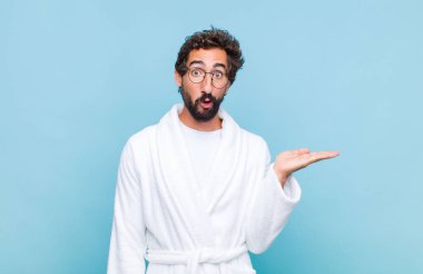 Banyo bornozu giymiş genç sakallı adam şaşırmış ve şoka uğramış görünüyor. Çenesi düşmüş ve eli açık bir objeyi tutuyor.