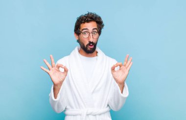 Banyo bornozu giyen genç sakallı adam şok olmuş, şaşırmış ve şaşırmış hissediyor. Onaylıyor. İki eliyle de onay işareti yapıyor.