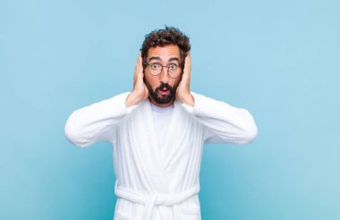 Banyo bornozu giymiş genç sakallı adam hoş olmayan bir şekilde şoka uğramış, korkmuş ya da endişeli görünüyor ağzı sonuna kadar açık ve iki kulağını da elleriyle kapatıyor.
