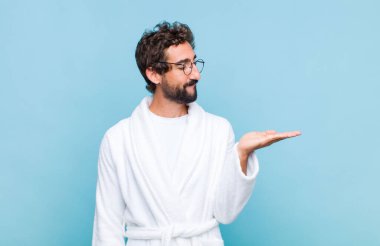 Banyo bornozu giymiş genç sakallı bir adam mutlu ve sıradan bir şekilde gülümsüyor, elinde tuttuğu bir nesneye ya da kavrama bakıyor.