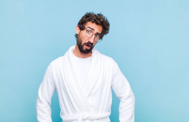 young bearded man wearing a bath robe feeling puzzled and confused, with a dumb, stunned expression looking at something unexpected