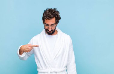 Banyo bornozu giymiş genç sakallı adam neşeyle gülümsüyor, yere bakıyor ve göğsüne işaret ediyor.