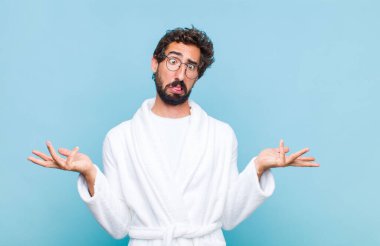 young bearded man wearing a bath robe shrugging with a dumb, crazy, confused, puzzled expression, feeling annoyed and clueless