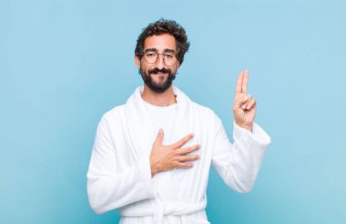 Banyo bornozu giyen genç sakallı adam mutlu, kendinden emin ve güvenilir görünüyor, gülümsüyor ve zafer işareti gösteriyor, olumlu bir tavırla.