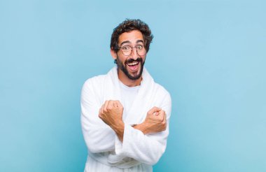 Banyo bornozu giymiş genç sakallı adam neşeli bir şekilde gülümsüyor ve kutluyor, yumruklarını sıkıp kollarını kavuşturuyor, mutlu ve pozitif hissediyor.