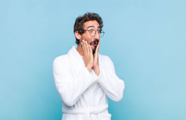 Banyo bornozu giyen genç sakallı adam mutlu, heyecanlı ve şaşırmış hissediyor, iki eli yüzü yana bakıyor