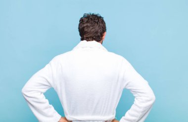 Banyo bornozu giymiş genç sakallı bir adam kafası karışık ya da dolu ya da şüphe ve soru dolu, merak ediyor, elleri kalçasında, dikiz aynasından