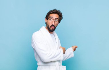 Banyo bornozu giymiş genç sakallı bir adam şaşkın ve şaşırmış hissediyor, yan taraftaki fotokopi alanını işaret ediyor, ağzı açık, şaşkın bir bakışla.