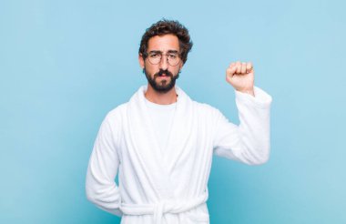 Banyo cübbesi giyen genç sakallı adam ciddi, güçlü ve asi hissediyor, yumruğunu kaldırıyor, protesto ediyor ya da devrim için savaşıyor.