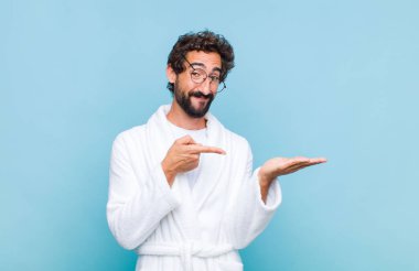 Banyo bornozu giymiş genç sakallı adam neşeyle gülümsüyor ve yan taraftaki fotokopi alanını işaret ederek bir nesneyi gösteriyor ya da reklamını yapıyor.