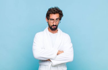 Banyo bornozu giyen genç sakallı adam hayal kırıklığına uğramış ve hayal kırıklığına uğramış görünüyor. Ciddi görünüyor, sinirli ve kollarını kavuşturmuş.