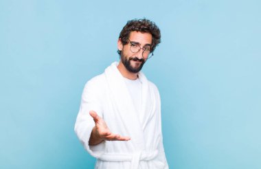 Banyo bornozu giymiş genç sakallı adam gülümsüyor, mutlu, kendinden emin ve arkadaş canlısı görünüyor, bir anlaşmaya varmak için el sıkışmayı teklif ediyor, işbirliği yapıyor.