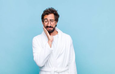 Genç sakallı bir adam banyo cübbesi giyiyor yanak tutuyor ve acı verici diş ağrısı çekiyor, hasta, mutsuz ve dişçi arıyor.