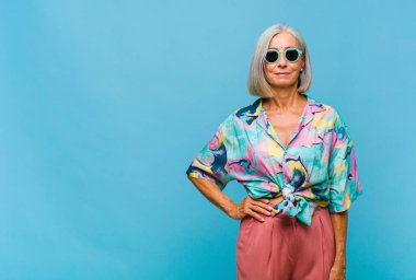 Orta yaşlı, havalı bir kadın mutlu bir şekilde gülümsüyor. Kendine güvenen, olumlu, gururlu ve arkadaş canlısı bir tavırla.