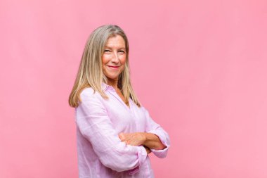 Orta yaşlı, güzel bir kadın kameraya çapraz kollarıyla gülümsüyor ve mutlu, kendinden emin, tatmin olmuş bir yüz ifadesi var.