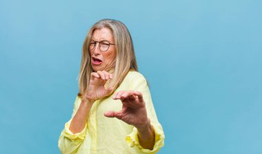 Orta yaşlı, güzel bir kadın panik ya da öfke içinde bağırıyor. Şok olmuş, korkmuş ya da öfkeli. Elleri başının yanında.