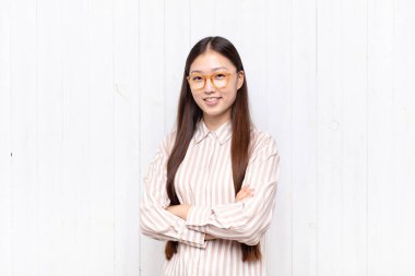 Asyalı genç bir kadın kameraya çapraz kollarla gülümsüyor ve mutlu, kendinden emin, tatmin olmuş bir ifade, yan görüş.