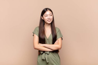 Asyalı genç bir kadın kendini mutlu, gururlu ve umutlu hissediyor, merak ediyor veya düşünüyor, çapraz kollarla fotokopi çekmeye çalışıyor.