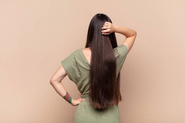 Asyalı genç kadın düşünüyor ya da şüphe ediyor, kafasını kaşıyor, kafası karışık ve kafası karışık hissediyor, arkaya ya da arkaya bakıyor.