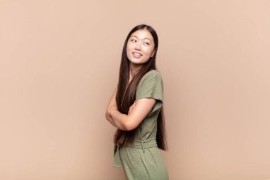 Asyalı genç bir kadın neşeyle gülümsüyor, mutlu, tatmin olmuş ve rahatlamış hissediyor, kollarını kavuşturup yan tarafa bakıyor.