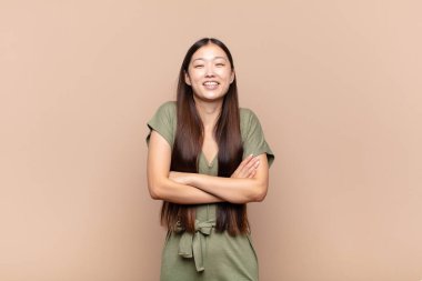 Asyalı genç bir kadın kollarını kavuşturup mutlu bir şekilde gülüyor, rahat, pozitif ve tatmin edici bir pozla.