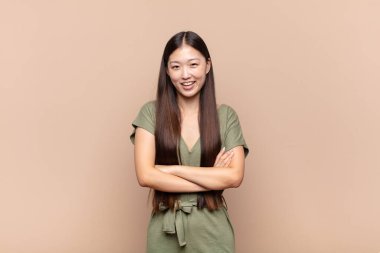 Asyalı genç bir kadın mutlu, gururlu ve tatmin olmuş görünüyor. Kollarını kavuşturup gülümsüyor.