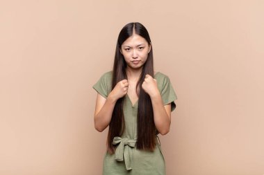 Asyalı genç kadın kendinden emin, sinirli, güçlü ve agresif görünüyor. Yumrukları boks pozisyonunda dövüşmeye hazır.