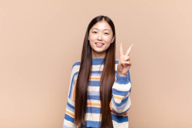 Asyalı genç kadın gülümsüyor ve arkadaş canlısı görünüyor. İki ya da iki numarayı gösteriyor. Elleri önde, geriye doğru sayıyor.