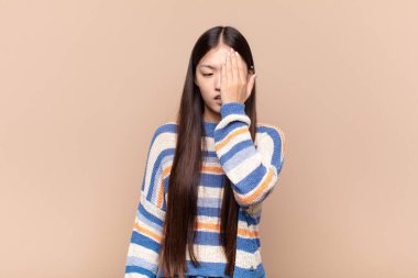 Asyalı genç bir kadın uykulu görünüyor, sıkılmış ve esniyor, baş ağrısı ve yüzünün yarısını kaplayan bir eliyle