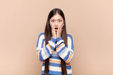 Asyalı genç kadın şok olmuş ve korkmuş hissediyor, ağzı açık ve elleri yanaklarında korkmuş görünüyor.