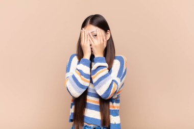 Asyalı genç bir kadın korkmuş ya da utanmış hissediyor. Gözleri yarı kapalı ya da elleri kapalı gözetliyor.