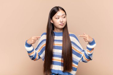 Asyalı genç bir kadın gururlu, pozitif ve rahat görünüyor. İki eliyle göğsünü gösteriyor.