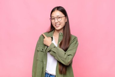 Asyalı genç kadın neşeyle gülümsüyor, mutlu hissediyor ve yan ve yukarı doğru işaret ediyor, fotokopi uzayındaki nesneleri gösteriyor